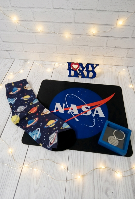 Conjunto de regalos de dia del padre sobre madera blanca con luces led, incluye calcetines de planetas, mosepad de la Nasa, llavero grabado y letrero "I love my dad" en azul con corazón rojo.
