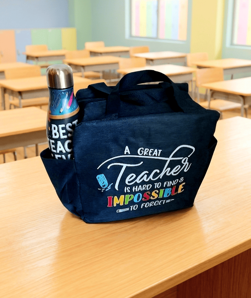 Primer plano de una bolsa termica de color azul marino con un diseño frontal colorido con la frase: "A Great Teacher is hard to find & Impossible to forget". En un bolsillo lateral puede verse parte de una botella termo. La bolsa con la botella se encuentran sobre una superficie de madera clara y un fondo de aula escolar.