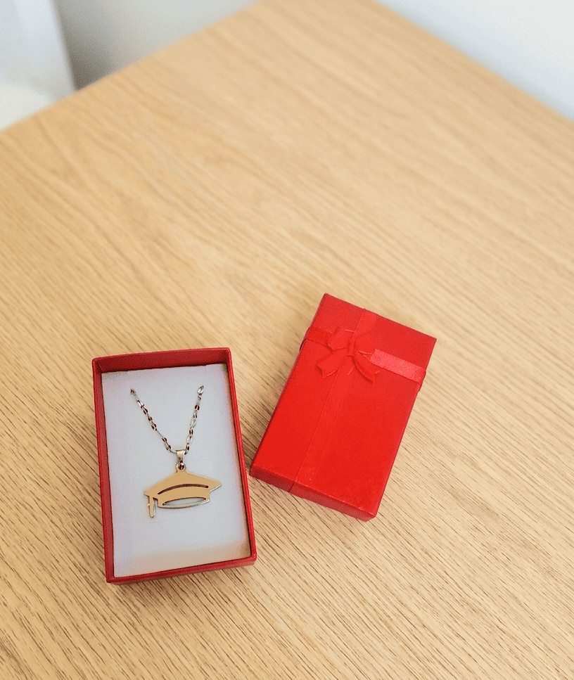 Primer plano de un colgante plateado de Acero inoxidable con un dije de birrete y cadena fina, presentado en una caja de joyeria roja con interior de color blanco, a su lado se encuentra la tapa de la caja de color rojo decorada con un lazo de raso rojo, todo se encuentra sobre una superficie de madera clara.