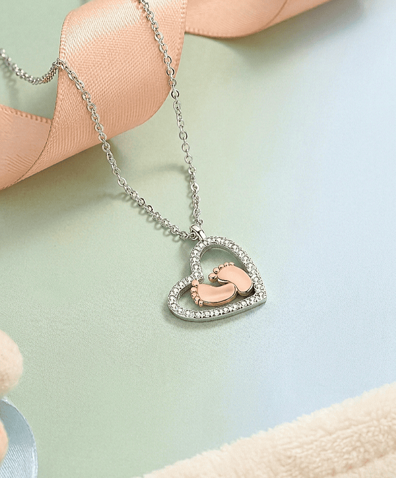 Colgante en forma de corazón con pies de bebé en su interior sobre una superficie suave en colores pastel.