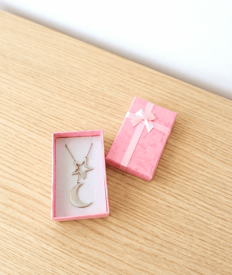 Colgante plateado de Acero inoxidable compuesto por una estrella y media luna entrelazados, colocado en el interior de una cajita de joyeria rosa sobre una superficie de madera clara.