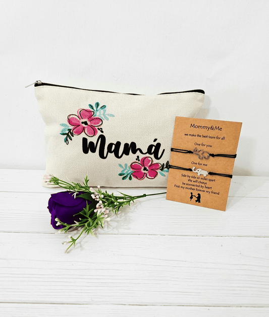 Primer plano de tres articulos de regalo sobre una superficie de madera blanca.A la izquierda un neceser de lona con la palabra "Mama".En el cento una rosa morada artificial. A la derecha una tarjeta kraft con dos pulseras de cuerda negra y dijes de elefantes plateados. La tarjeta lleva el título de" Mommy & me" y contiene un texto en ingles.