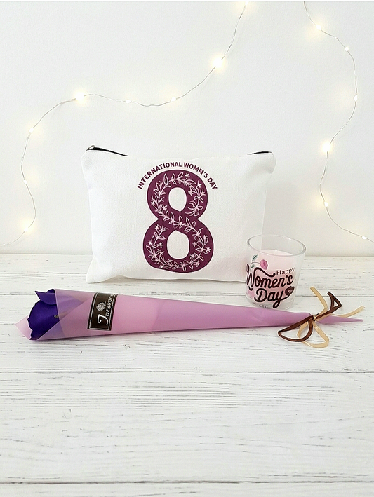 Neceser blanco con numero 8 en color morado junto a una vela rosa con la frase " Happy women's day" y una rosa de color morado con envoltorio en color malva, todo junto sobre una superficie de madera blanca y luces led de fondo.