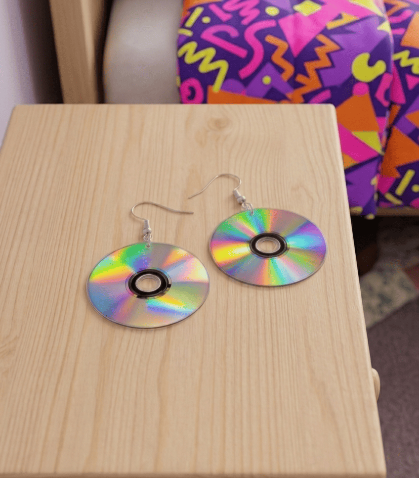 Pendientes con forma de CD sobre una superficie de mesita de noche con un fondo de habitación en el que se ve una cama cubierta con un edredón de dibujos assimetricos en colores fluor.