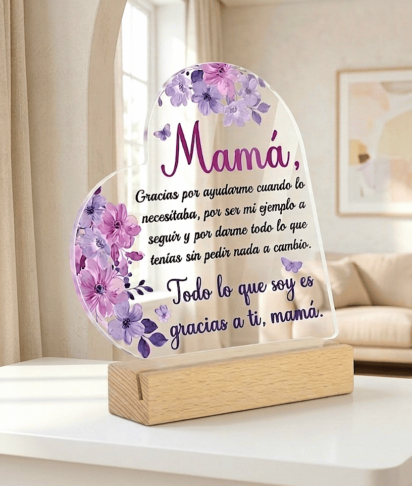 Placa decorativa de metacrilato en forma de corazón con un texto emotivo de dia de la madre decorado con flores moradas sobre una base de madera clara en una mesa blanca con un fondo acogedor de salón.