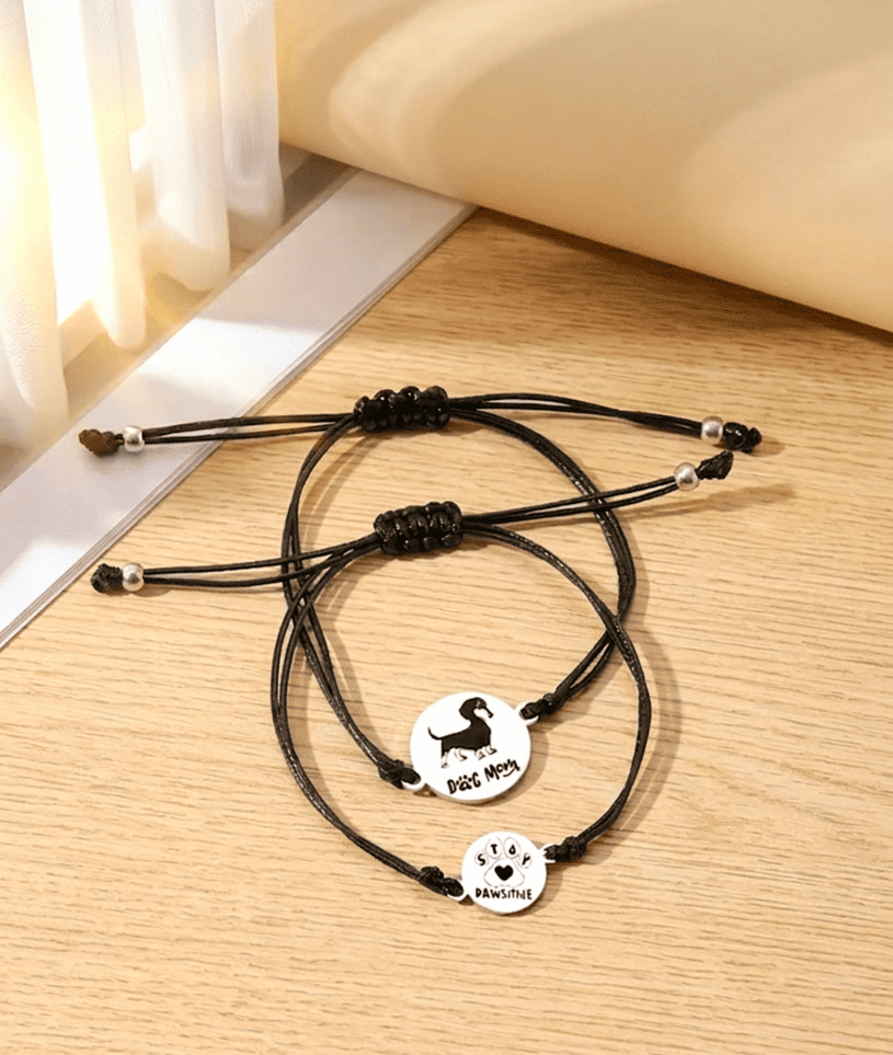Vista superior de dos pulseras de cordón negro ajustables con dijes circulares de Acero Inoxidable con detalles de perro y huella de pata sobre superficie de madera clara.