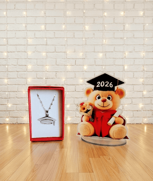 Conjunto de regalos de Graduación formado por un colgante plateado de Acero Inoxidable con dije de birrete presentado en caja de joyeria roja con interior blanco, al lado puede verse una figura acrilica de osito vestido de graduación con un ramo de flores y un diploma, sobre una base acrílica ovalada, en su birrete puede verse el año 2026. Los dos regalos pueden verse sobre una superficie de madera clara y una pared blanca con luces led.