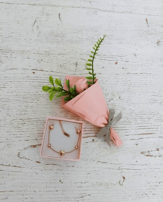 Sobre una superficie de madera blanca se puede ver una caja de joyeria con una pulsera dorada en Acero con corazones y cristales brillantes, a su lado una Rosa Eterna con envoltorio satinado de color rosa con lazo gris decorativo.