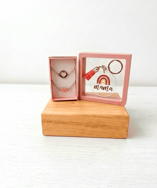 Set de regalos compuesto por dos pulseras de Acero inoxidable con dijes de corazón junto a llavero circular de metacrilato con diseño de arcoiris en tonos rosa y la palabra Mamá sobre una base de madera clara.