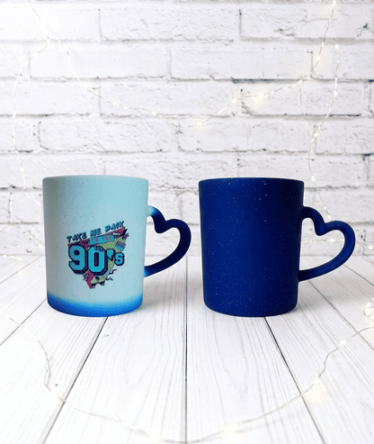 Tazas de cerámica termosensibles, la taza de la izquierda contiene bebida caliente y puede verse un diseño de los 90 con colores fluor, la taza de la derecha no contiene bebida y puede verse su color original azul cobalto con efecto brillante, están sobre una superficie de madera blanca, pared de ladrillo visto y luces led.