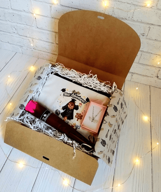 Caja abierta de regalo vista desde arriba con un neceser color crudo con un diseño de graduación con frase motivadora en inglés, sobre el neceser se puede ver una cajita de joyeria con un colgante en Acero inoxidable de corazón con alas brillantes, en la parte inferior se encuentra una Rosa Eterna con envoltorio satinado de color Borgoña, la caja se encuentra sobre una superficie de madera blanca y luces led.