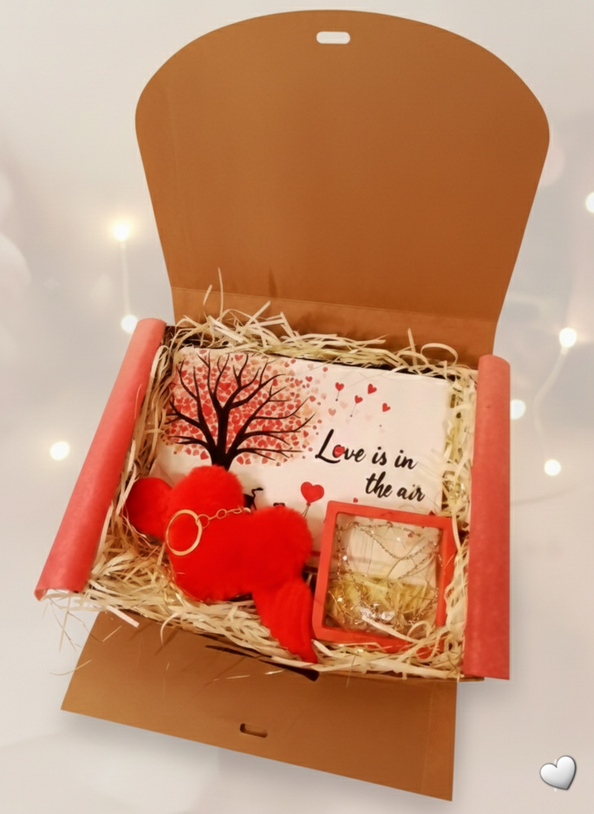 Caja de regalo abierta, con eceser de corazones, colgante de corazon con alas y llavero rojo de peluche con alas.