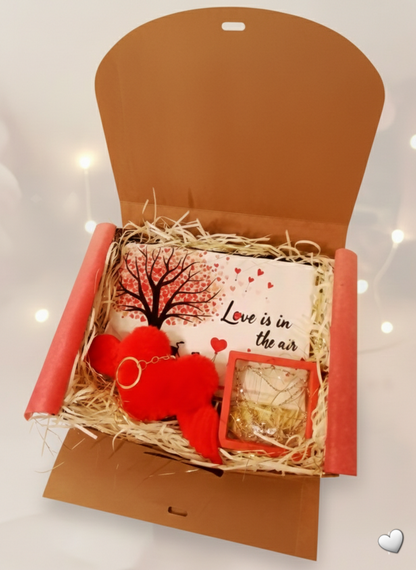 Caja de regalo abierta, con eceser de corazones, colgante de corazon con alas y llavero rojo de peluche con alas.