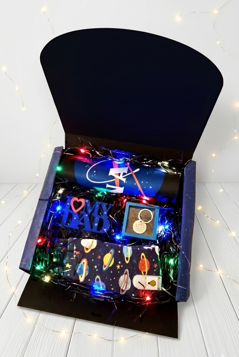 Caja de regalo negra abierta contiene calcetines con diseño de planetas, llavero en caja transparente con marco azul y mousepad de la Nasa enrrollado sobre virutas de papel y luces led de colores 