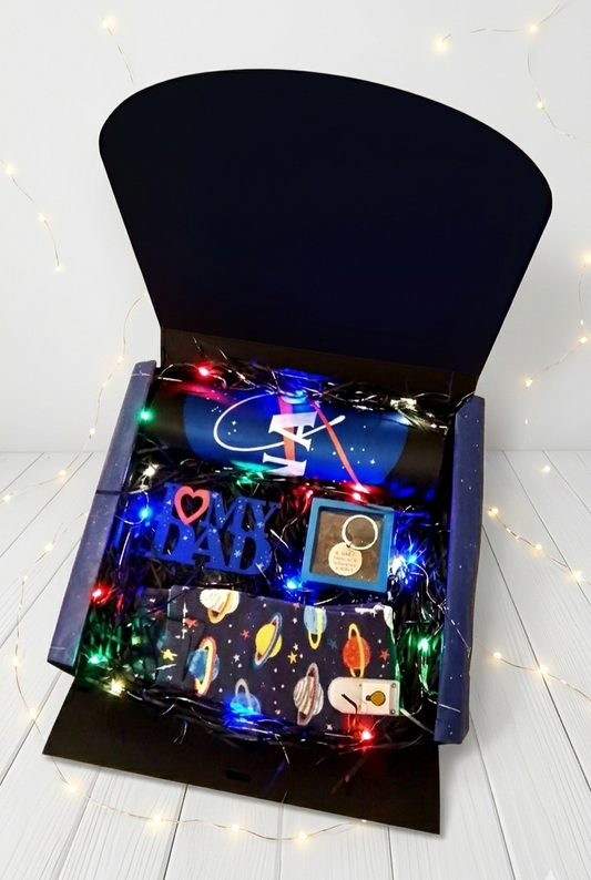 Caja de regalo negra abierta contiene calcetines con diseño de planetas, llavero en caja transparente con marco azul y mousepad de la Nasa enrrollado sobre virutas de papel y luces led de colores 