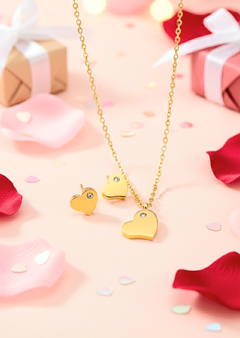 Colgante y pendientes dorados de Acero en forma de corazón con un pequeño cristal brillante, presentados sobre un fondo rosa con petalos.