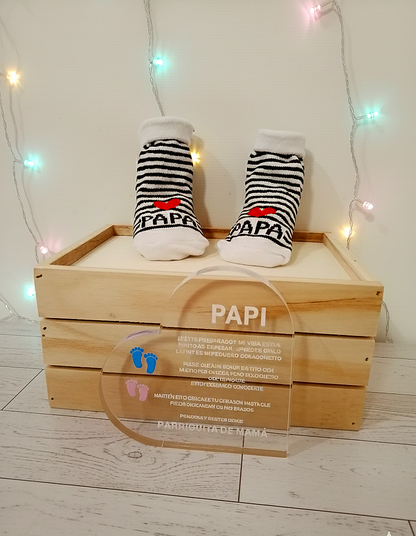 Conjunto de articulos de regalo de dia del padre compuesto por una placa de metacrilato transaparente con texto en español de dia del padre y calcenines de bebé sobre caja de madera y fondo de luces led de colores.
