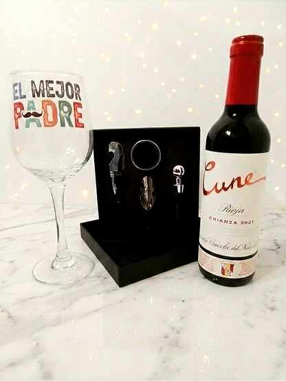 Primer plano de una botella de vino Cune Rioja junto a una copa personalizada y un kit de accesorios de vino en caja negra sobre fondo de mesa en marmol blanco y luces led decorativas.