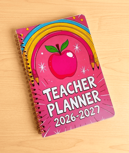 Primer plano vista desde arriba de una agenda de color fucsia con diseño  Retro-Pop con un arcoiris de lapiceros y una manzana, debajo puede leerse TEACHER PLANNER BOX 2026-27 con tipografia clásica de color blanca sobre una superficie de madera clara. 
