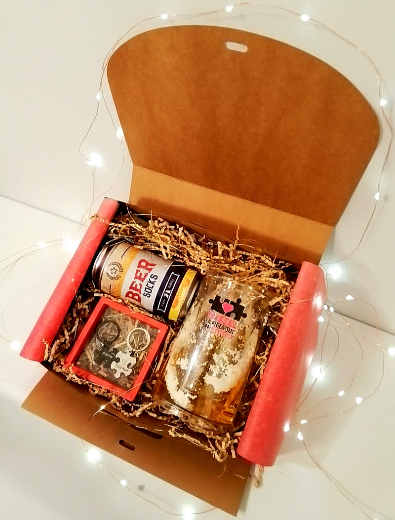 Caja de regalo abierta Beerly Fit Box, mostrando calcetines en lata, una jarra de cerveza personalizada y llaveros de piezas de puzzle sobre virutas de papel decorativas con luces led de fondo.