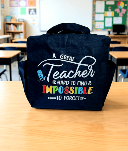 Primer plano de una bolsa termica de color azul marino con asas y cierre de cremallera con dos bolsillos laterales. En la parte frontal puede leerse la frase : "A Great Teacher is hard to find & Impossible to forget" con varios tipos de tipografia en color blanco y multicolor sobre superficie de madera clara y un fondo de aula escolar.