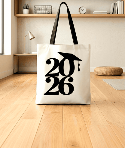 Bolsa Tote en color blanco con numeros de color negro formando 2026 con un detalle de la silueta de un birrete sobre una superficie de madera clara y un fondo de habitacion juvenil.
