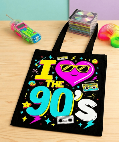 Bolsa tote negra con diseño de los 90 con la frase "I love the 90's" y detalles de cintas de caset y estrellas de colores fluor sobre una superficie de madera clara y accesorios de los 90.