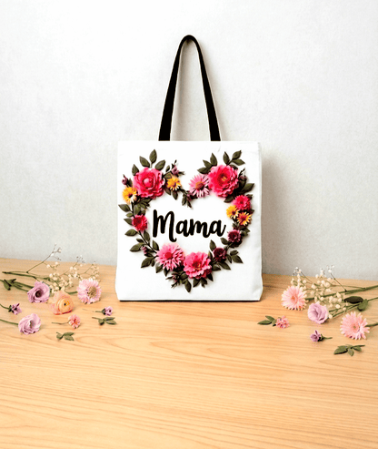 Bolsa Tote de lona balnca con la palabra Mama con letras negras y flores de varios colores formando un corazón alrededor,  sobre una superficie de madera clara con varias flores en tonos pastel a los dos lados.