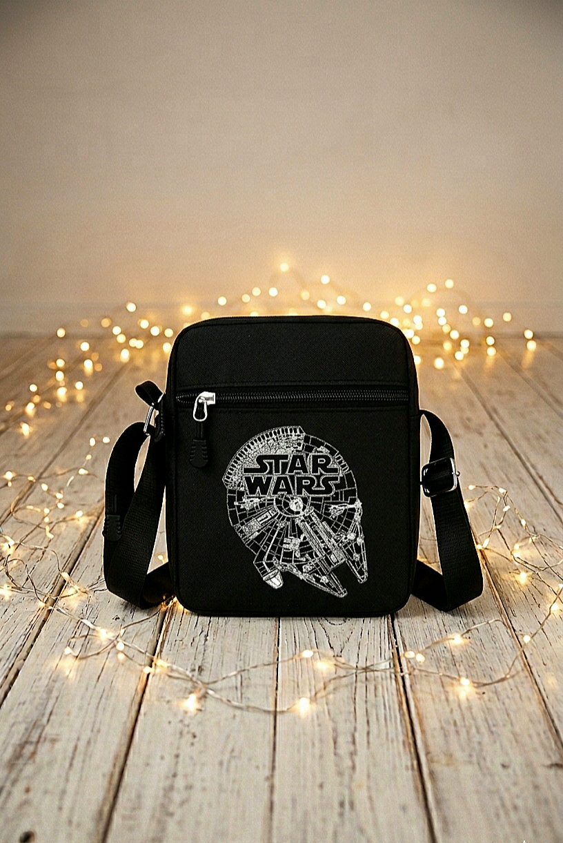 Un bolso bandolera negro con diseño en blanco de Millennium Falcon y el logo de Star Wars rodeado de luces led sobre una superficie de madera blanca.