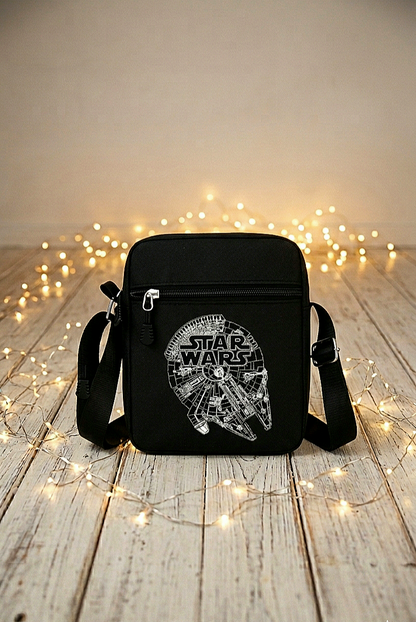 Un bolso bandolera negro con diseño en blanco de Millennium Falcon y el logo de Star Wars rodeado de luces led sobre una superficie de madera blanca.