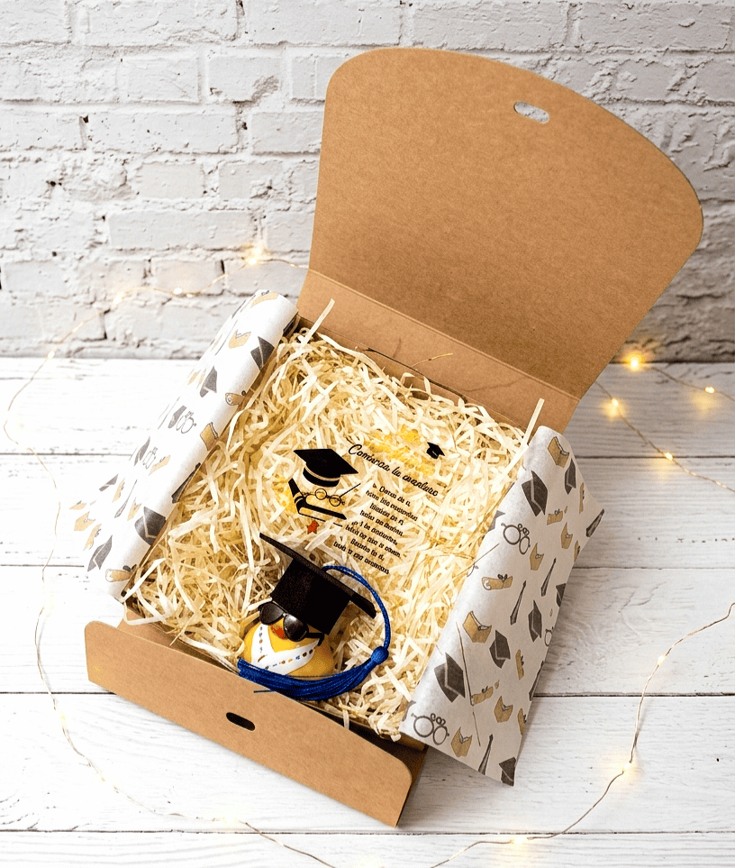Caja de regalo abierta vista desde arriba, contiene una placa con un texto emotivo de graduación, en la parte inferior hay un patito de goma equipado con un birrete con borla azul, unas gafas de sol y una estola de graduación con letras de colores todo sobre una superficie de madera blanca con luces led.