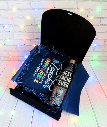 Caja de regalo abierta vista desde arriba, mostrando una bolsa termica plegada con un texto de agradecimiento en inglés, con dos tipos de tipografia: Una tipografia moderna en color blanco y otra tipografia mas clásica y con letras de colores, a su lado hay una botella termica con interior de acero inoxidable y se puede leer la frase "Best Teacher Ever" con ilustraciones y motivos escolares. La caja se encuentra sobre una superficie de tarima de madera blanca con luces led de colores.