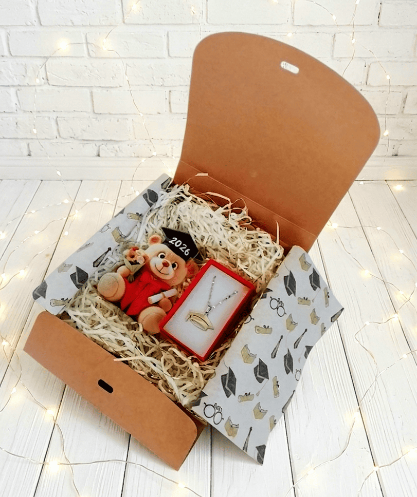 Caja de regalo abierta vista desde arriba,  mostrando una figura acrilica de osito con birrete y toga de graduacion con el año 2026 en el birrete y un diploma en la mano, a su lado puede verse una cajita de joyería con un colgante de Acero Inoxidable con un dije de birrete, la caja se encuentra sobre una superficie de tarima de madera blanca y luces led.