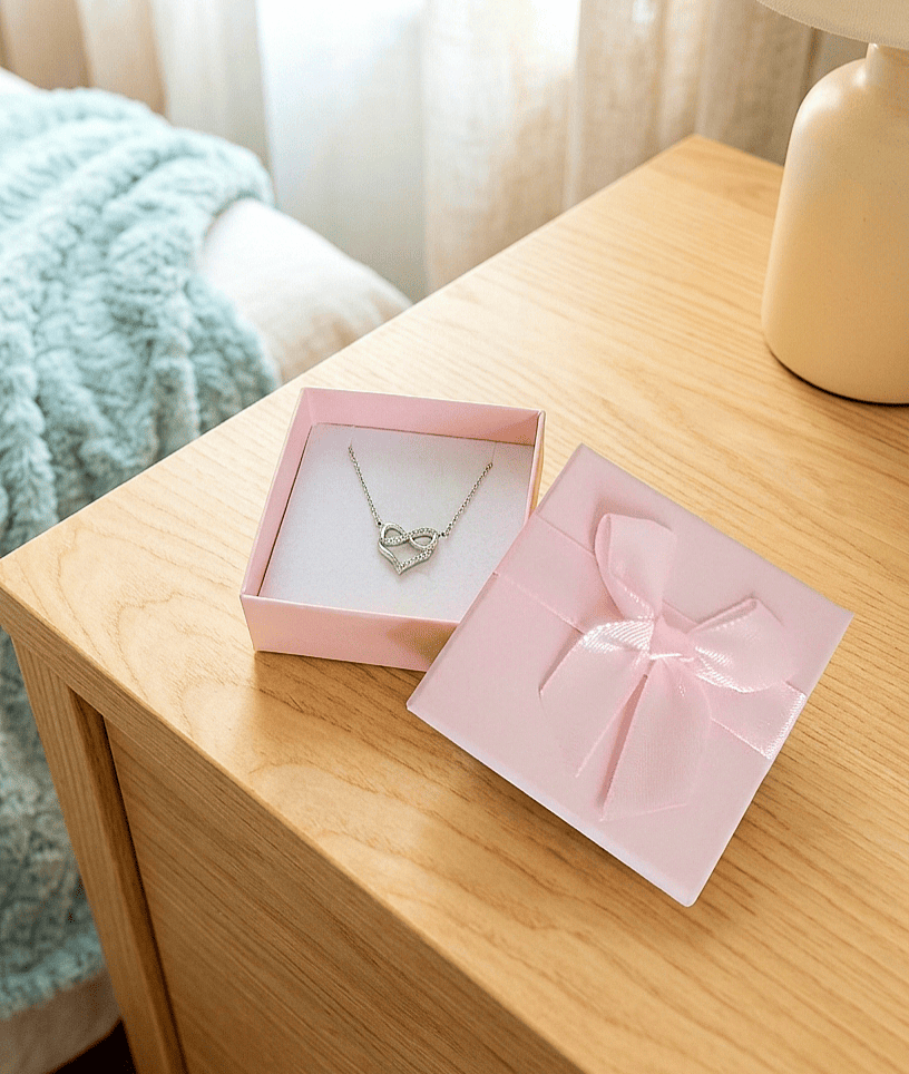 Una caja de joyería abierta con la tapa apoyada, de color rosa con un lazo de raso en color pastel, en su interior se puede ver un colgante plateado con un corazón entrelazando el simbolo de infinito sobre una mesita de noche de madera clara y un fondo de dormitorio en colores pastel.