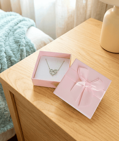Una caja de joyería abierta con la tapa apoyada, de color rosa con un lazo de raso en color pastel, en su interior se puede ver un colgante plateado con un corazón entrelazando el simbolo de infinito sobre una mesita de noche de madera clara y un fondo de dormitorio en colores pastel.