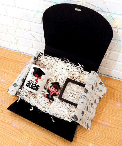 Caja negra de regalo abierta vista desde arriba que contiene una taza de cerámica con una ilustración de un birrete con un diploma, debajo puede leerse la frase "Class of 2026" a su lado puede verse una figura acrilica que representa a un graduado vestido con toga y birrete mostrando una expresión de gran alegría, por ultimo hay un llavero redondo con una frase de felicitación presentado en caja de exhibición con marco negro, todo sobre virutas de papel blanco en superficie de madera clara y luces led.