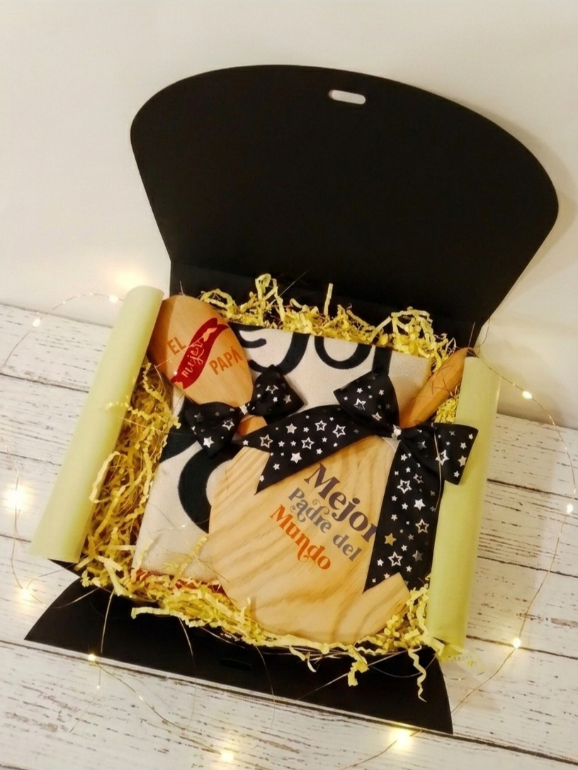 Caja de regalo negra abierta que contiene una cuchara de madera grabada , una mini tabla de cortar, un delantal plegado decorado con dos lazos negros con estrellitas plateadas, todo sobre virutas de papel amarillas en fondo de madera blanca y luces led