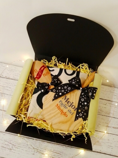 Caja de regalo negra abierta que contiene una cuchara de madera grabada , una mini tabla de cortar, un delantal plegado decorado con dos lazos negros con estrellitas plateadas, todo sobre virutas de papel amarillas en fondo de madera blanca y luces led