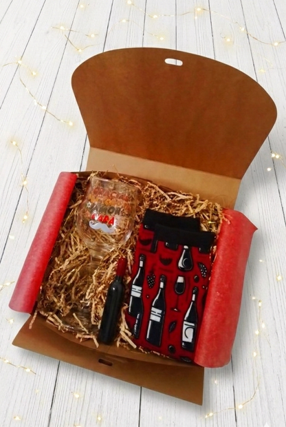 Caja de regalo abierta que incluye una copa de vino personalizada, calcetines color burdeos con estampado de botellas de vino y copas y sacacorchos en forma de botella de vino sobre fondo de madera blanca con luces led.