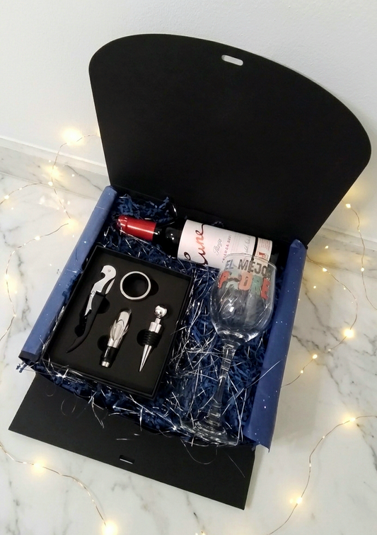 Vista superior de la experiencia unboxing de WINE MASTER BOX: Una caja de regalo negra con relleno de virutas de papel azul que contiene una botella de vino Cune Rioja, una copa "El Mejor Padre" y un set de accesorios de acero sobre fondo de marmol con luces led decorativas.