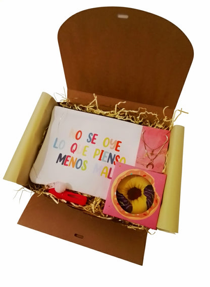 Caja de regalo abierta, mostrando un neceser con letras multicolor, un colgante con luna creciente, calcetines formando un Donuts y atomizador con detalle de pompón, con decoracion de virutas de papel y papel de seda amarillo
