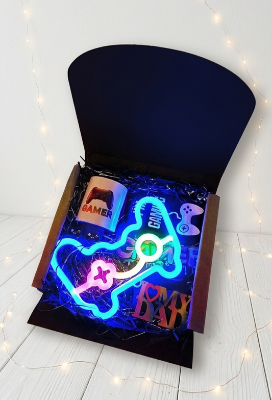 Caja de regalo negra abierta contiene lampara led con forma de mando de consola, calcetines negros con letras blancas, taza bicolor con diseño de mando de consola, protectores de cables y letrero de colores fluor I love my dad  sobre superficie de madera blanca con luces led
