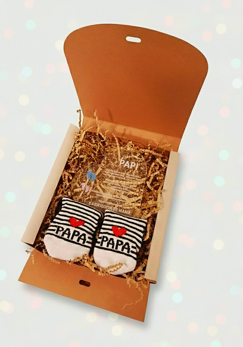 Caja abierta mostrando una placa de metacrilato con forma de corazón y frase emotiva de dia del padre y calcetines de bebé de rayas sobre virutas de papel Kraft. 