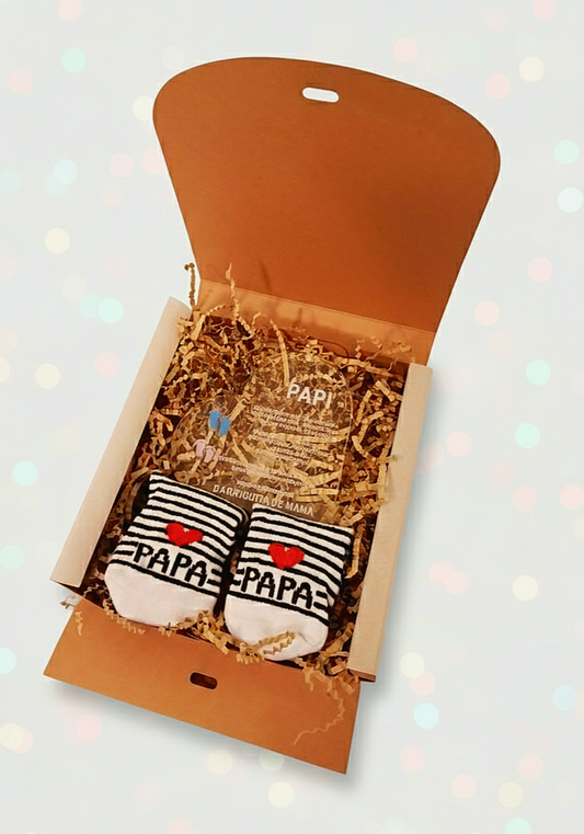 Caja abierta mostrando una placa de metacrilato con forma de corazón y frase emotiva de dia del padre y calcetines de bebé de rayas sobre virutas de papel Kraft. 