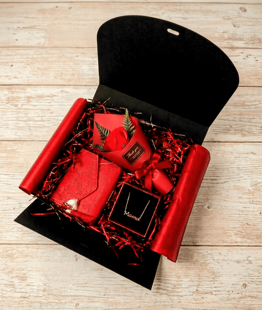 Vista desde arriba de una caja de regalo negra abierta que contiene un monedero rojo, un colgante dorado con la palabra Mamá y una Rosa Eterna de jabón de color roja con relleno decorativo sobre superficie de madera clara.