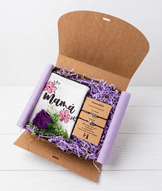 Vista superior de la caja de abierta para el dia de la madre, llamada Mommy & me box. Contiene un neceser de lona color blanco con la palabra Mama y dos flores de loto. A su lado, una tarjeta kraft  con dos pulseras de cuerda negra con dijes de elefantes plateados. Completa el conjunto una rosa morada todo sobre una superficie de madera blanca.