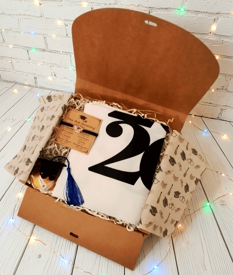 Caja de regalo abierta vista desde arriba que contiene una bolsa tote plegada de color blanca con grandes números en color negro, en la parte superior izquierda puede verse una pulsera de cordón negro con dije metalico en forma de birrete presentada en tarjeta kraft con emotivo texto en inglés, en la parte inferior hay un pato de goma con birrete de borla azul, gafas de sol y estola de graduación. Todo sobre una superficie de madera blanca con lucecitas led de colores.