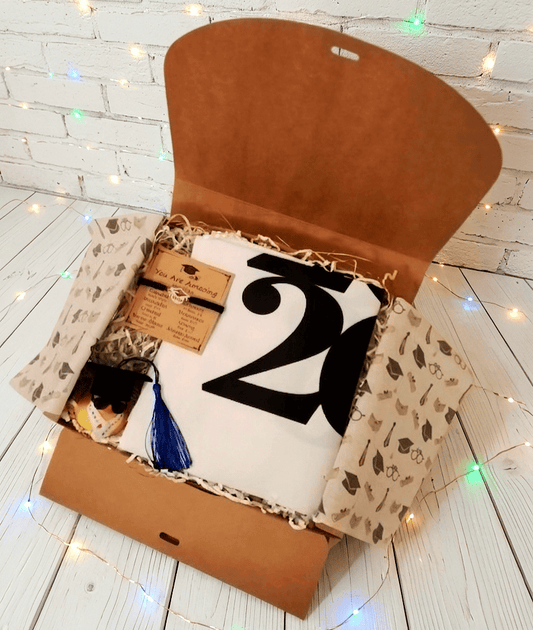 Caja de regalo abierta vista desde arriba que contiene una bolsa tote plegada de color blanca con grandes números en color negro, en la parte superior izquierda puede verse una pulsera de cordón negro con dije metalico en forma de birrete presentada en tarjeta kraft con emotivo texto en inglés, en la parte inferior hay un pato de goma con birrete de borla azul, gafas de sol y estola de graduación. Todo sobre una superficie de madera blanca con lucecitas led de colores.