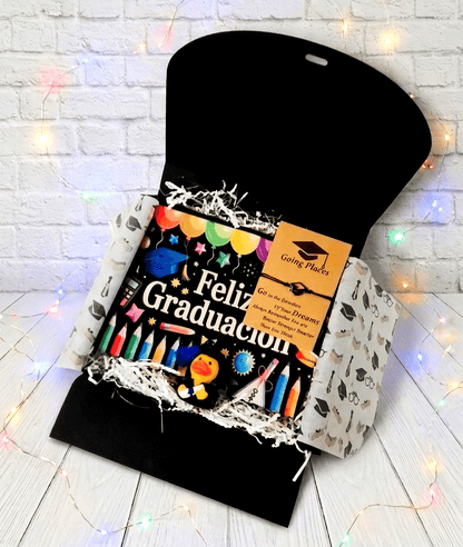 Caja de regalo abierta vista desde arriba, en su interior puede verse una bolsa multiusos de color negro con un diseño de dibujos alegres multicolores con el texto en letras blancas "FELIZ GRADUACIÓN". En la parte superior izquierda hay una pulsera de cordón negro con dije de birrete, presentada en tarjeta de papel kraft con un texto motivador en inglés. De fondo una superficie blanca con pequeñas luces led en colores pastel.
