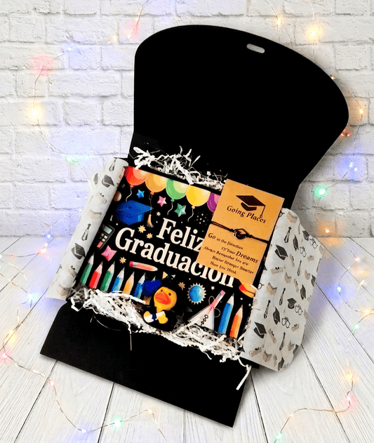 Caja de regalo abierta vista desde arriba, en su interior puede verse una bolsa multiusos de color negro con un diseño de dibujos alegres multicolores con el texto en letras blancas "FELIZ GRADUACIÓN". En la parte superior izquierda hay una pulsera de cordón negro con dije de birrete, presentada en tarjeta de papel kraft con un texto motivador en inglés. De fondo una superficie blanca con pequeñas luces led en colores pastel.